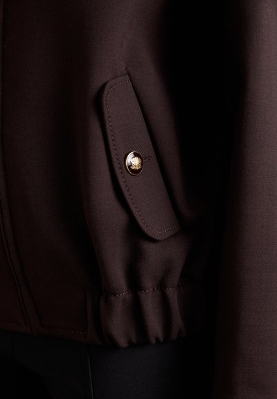 Braune Jacke mit strukturiertem Stoff, einer Klappentasche mit einem goldenen Knopf und gerafftem Saumdetail. Nahaufnahme des Ärmels.