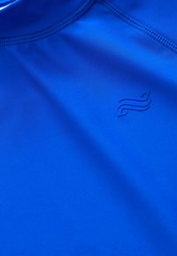 Zils sporta krekliņš, kas izgatavots no gludas, elastīgas auduma, ar apļveida kakla izgriezumu. Tam ir uzkrītoša logo krūšu tuvumā un plakanas šuves.