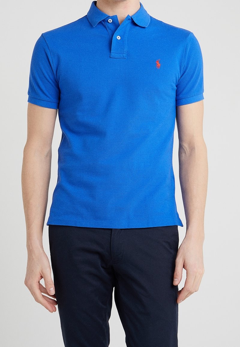 Homme portant un polo à manches courtes bleu vif avec un petit logo rouge sur la poitrine et un pantalon foncé.