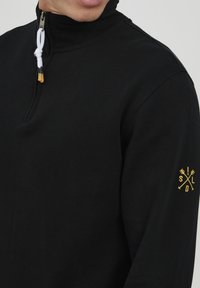 Pullover nero con colletto alto, dotato di mezza zip e coulisse bianca. La manica sinistra presenta un accento con logo dorato. Materiale in cotone morbido.