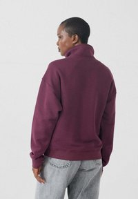 Pullover bordeaux realizzato in morbido materiale con colletto alto, spalle scese e polsini a coste, abbinato a jeans grigio chiaro.