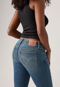 Jean skinny en denim bleu moyen avec une finition délavée, présentant un patch en cuir, deux poches arrière et un détail de couture caractéristique.