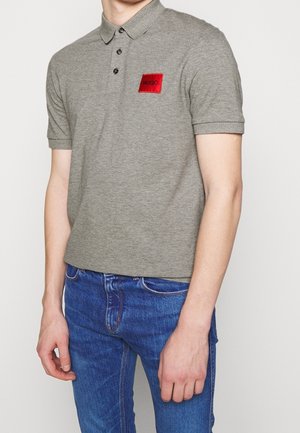 Polo gris avec une finition texturée, arborant un patch rectangulaire rouge portant l'inscription "HUGO". Manches courtes et col classique.