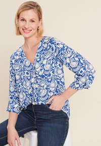 Blusa con stampa floreale blu e motivi bianchi, scollatura a V e maniche a tre quarti, abbinata a jeans di denim scuro.
