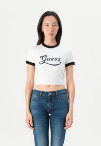 Hvit kortermet t-skjorte med svart ringkrage, med en svart skriptlogo "Guess" over brystet, kombinert med blå denimjeans.
