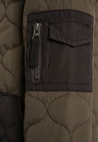 Kurtka pikowana w oliwkowym zielonym kolorze z ciemnoszarymi akcentami. Posiada kieszeń na zamek oraz kieszeń z klapką na ramieniu.