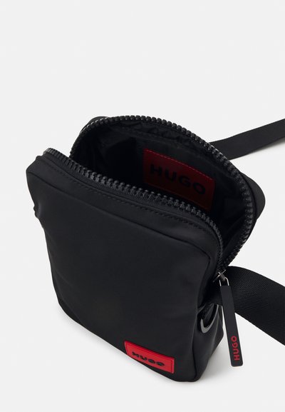 Sac en nylon noir avec fermeture éclair, doté d'un patch logo rouge, d'une bandoulière réglable et d'une texture lisse. L'intérieur présente une étiquette de marque.
