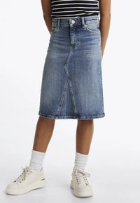 Gonna di jeans con cucitura frontale, taglio a ginocchio, colore blu chiaro e leggere sfumature. Abbinata a sneakers bianche e calzini a coste.