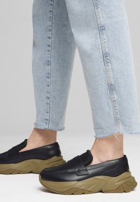 Pantofi negri tip slip-on cu o talpă groasă de culoare bej; prezintă un exterior din piele netedă și accente elastice. Purtați cu blugi scurți de culoare albastru deschis.
