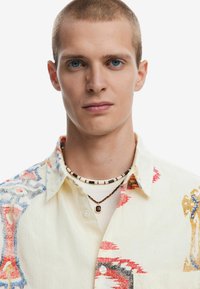 Camicia a maniche corte color crema con motivi astratti colorati; presenta una chiusura con bottoni e un colletto testurizzato; abbinata a collane di perline.