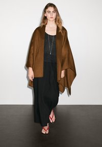 Brun ullponcho draperad över en svart topp och lång kjol. Röda flip-flops fullföljer outfiten. Slät struktur, lös design.
