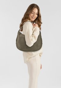 Olivgrüne Leder-Schultertasche mit runder Form, einem Schulterriemen und dezentem Markenlogo. Das Modell trägt einen cremigen Pullover und eine Hose.