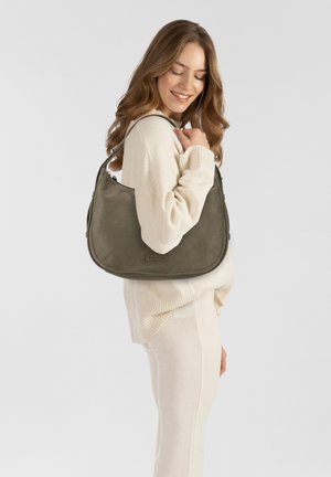 Olivgrüne Leder-Schultertasche mit runder Form, einem Schulterriemen und dezentem Markenlogo. Das Modell trägt einen cremigen Pullover und eine Hose.