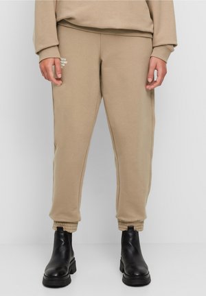 Person iført beige joggerbukser med lille hvidt logo på venstre lår og sorte ankellange støvler mod en ensfarvet baggrund.