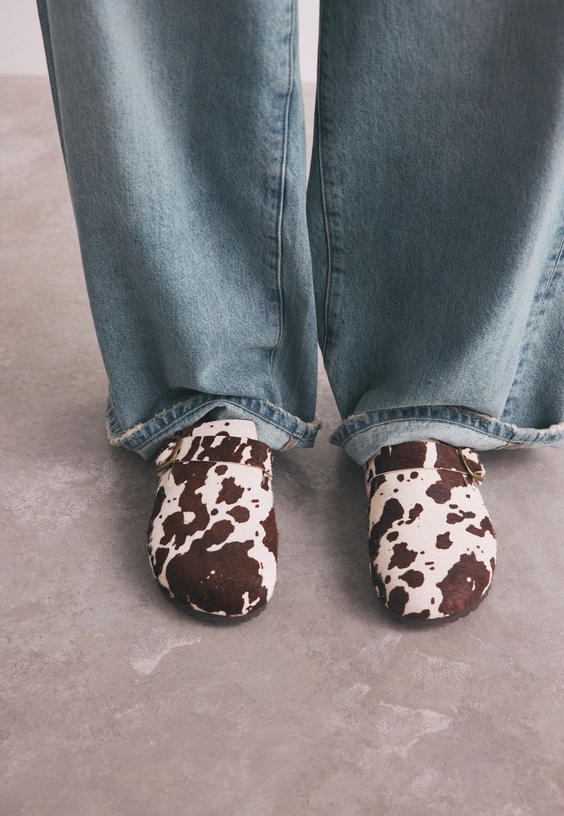 Personne portant un jean en denim bleu clair et des chaussures slip-on à imprimé vache, debout sur un sol gris.
