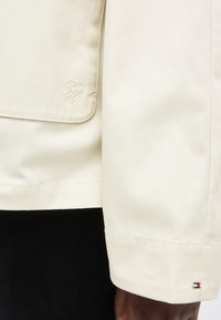 Μανίκι και λεπτομέρεια τσέπης σε κρεμ χρώμα, με διακριτικά κεντημένα και ραμμένα λογότυπα, φορεμένα πάνω από μαύρα ρούχα.