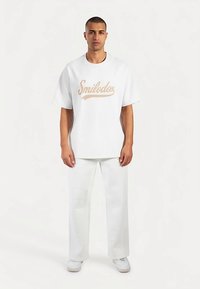 Wit oversized T-shirt met het "Smilodox" logo in lichtbruin. Gecombineerd met witte wijde broekspijpen en witte sneakers. Soepel textuur.