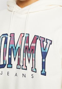 Krémová mikina s výrazným modro-růžovým károvaným logem "TOMMY". Obsahuje bílé šňůrky na stažení a černé okraje kolem písmen.