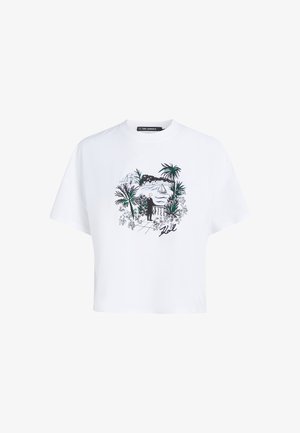 T-shirt blanc avec un dessin noir et vert représentant une personne debout près d'une balustrade surplombant l'eau, un voilier, des palmiers et des bâtiments au loin.