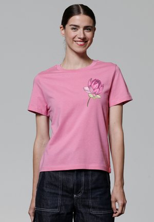 T-shirt en coton rose à manches courtes, col rond, avec un graphisme de fleur de lotus sur le côté gauche de la poitrine. Texture douce, coupe décontractée.