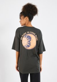 Donkergroene oversized t-shirt met een cirkelvormige grafiek van een zon en maan ontwerp, vergezeld van de tekst "WEES VRIENDELIJK VOOR JE GEEST."