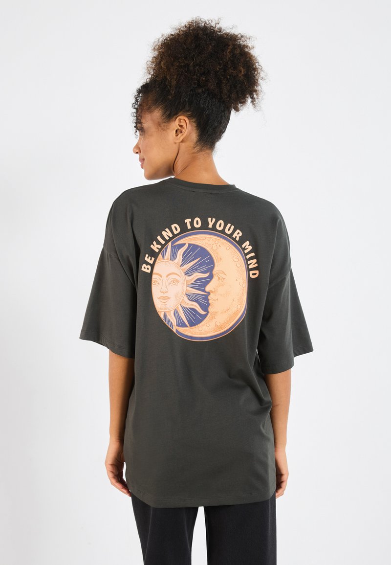 Donkergroene oversized t-shirt met een cirkelvormige grafiek van een zon en maan ontwerp, vergezeld van de tekst "WEES VRIENDELIJK VOOR JE GEEST."