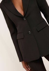 Fekete szabott blézer mély V-nyakkivágással, strukturált vállakkal, két elülső zsebbel és egy gombos záródással. Simaszerű anyag, matt felülettel.