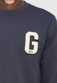 Mørkeblå hettegenser med en teksturert hvit "G" lapp med svart kant, og ordet "Grant" sydd nedenfor. Mykt bomullsmateriale.
