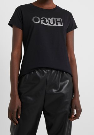 Musta puuvillainen T-paita, jossa on kiiltävä hopeinen "HUGO"-logo rinnassa. Lyhyet hihat ja rento istuvuus. Yhdistetty kiiltäviin mustiin housuihin.