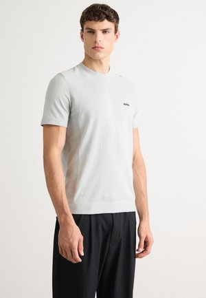 Un homme portant un t-shirt tricoté à manches courtes gris clair avec le logo "BOSS" sur la poitrine et un pantalon noir, se tenant devant un fond blanc uni.