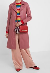 Vaaleanpunainen nahkainen trenssitakki, jossa kaulus, yhdistetty moniväriseen raidalliseen neulepaitaan, satiiniset leveälahkeiset housut ja punaiset asusteet.