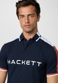 Marineblauwe polo met contrasterende witte en roestkleurige accenten, met een logo "HACKETT" in vette witte letters op de borst en een klassiek kragen.