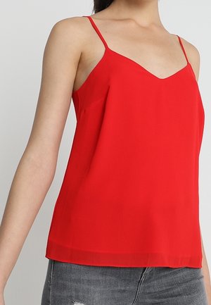 Kobieta w czerwonym topie typu camisole bez rękawów z cienkimi ramiączkami, dopasowanym do szarych, postrzępionych jeansów na jednolitym tle.