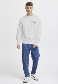 Lysebeige genser med grønn brodering, rund hals og lange ermer. Kombinert med løse blå jeans og svart-hvite sneakers.