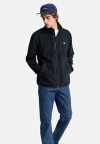 Giacca in pile nera con colletto alto, chiusura con zip, tasca sul petto e due tasche laterali. Abbinata a jeans in denim blu.