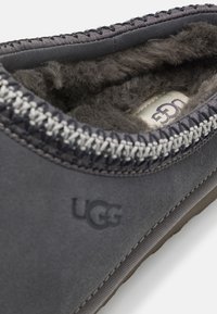 Scarpa UGG di colore grigio scuro con suola in gomma piatta, tomaia in suede testurizzato, cuciture grigie e una morbida fodera in pelliccia visibile all'interno. Logo UGG impresso.