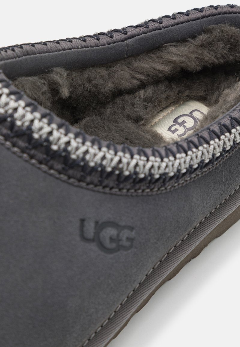 Dunkelgrauer UGG-Schuh mit flacher Gummisohle, strukturiertem Wildleder-Oberteil, grauen Nähten und weichem Fellfutter, das innen sichtbar ist. UGG-Logo geprägt.