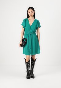 Robe turquoise avec décolleté en V et manches volantes, cintrée à la taille. Bottes de cowboy noires jusqu'aux genoux avec des coutures blanches intriquées. Tenant un sac noir.
