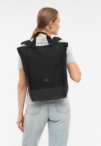 Fekete hátizsák strukturált dizájnnal, szövet anyagból, elülső cipzáras zsebbel és rövid fogantyúval, elülső logóval ellátott folttal.