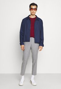 Marineblauer Reißverschlussjacke, burgunderfarbenes Rundhals-Shirt, hellgraue Slim-Fit-Hosen. Weiße Sneakers und rotgetönte Brille runden das Outfit ab.