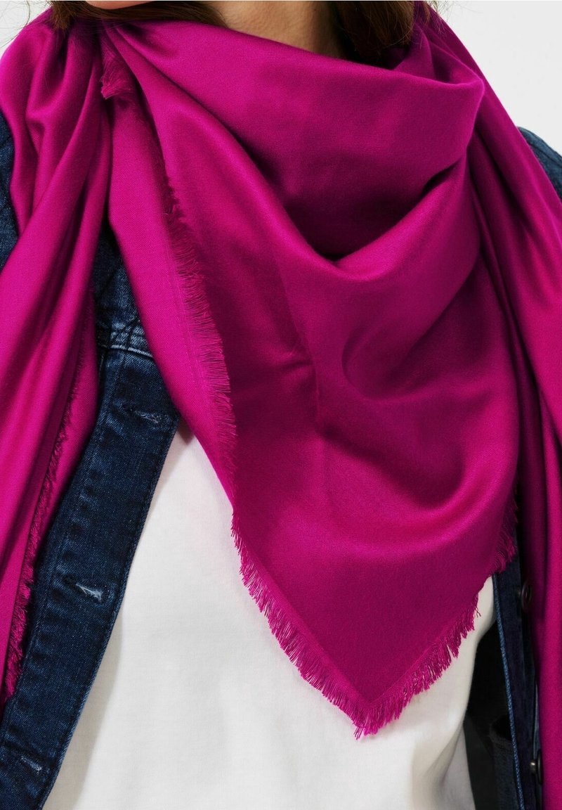 Écharpe magenta vif aux bords frangés drapée sur une chemise blanche et une veste en denim foncé portée par une personne.