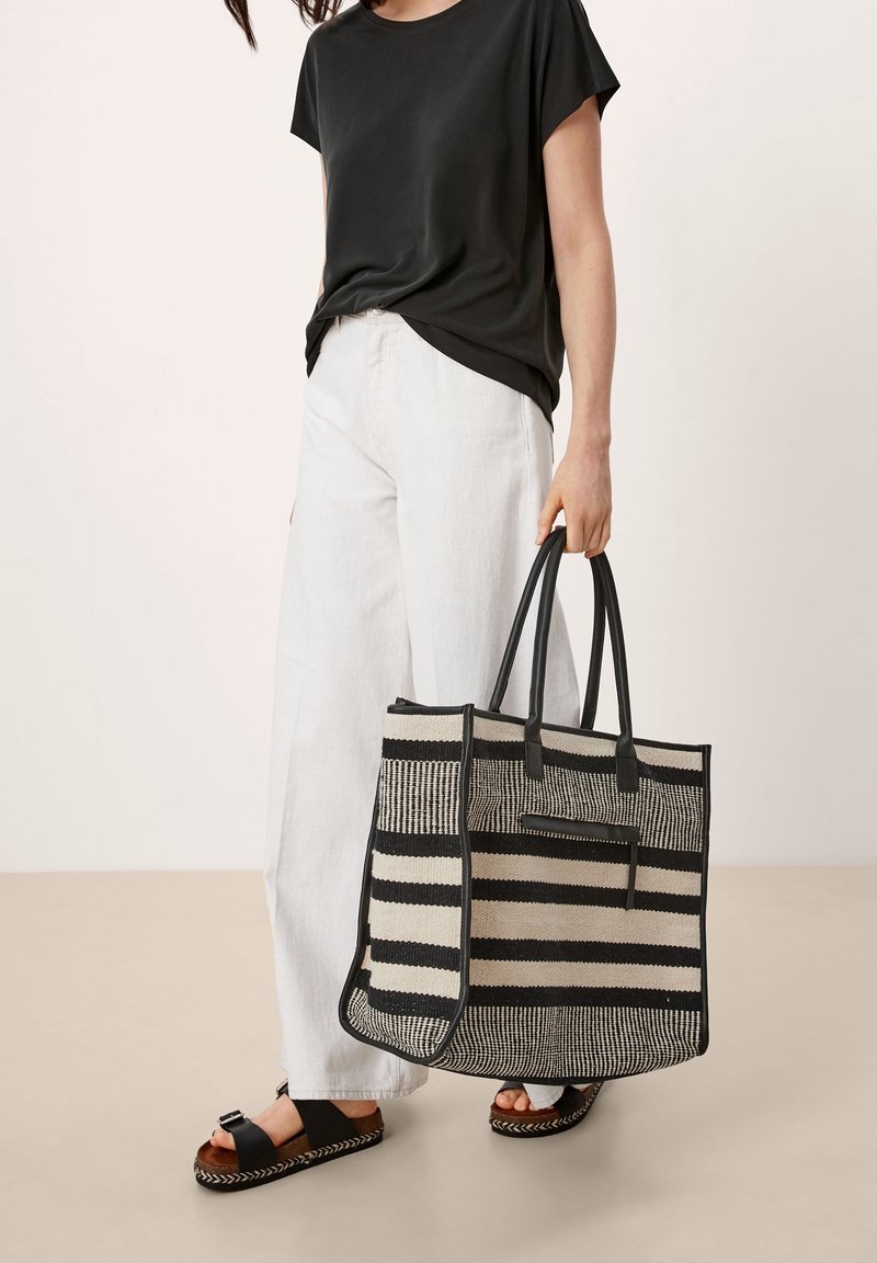 Sac fourre-tout tissé avec des rayures noires et crème, de forme rectangulaire, poignées en cuir et poche zippée. Associé à un pantalon clair et des sandales.