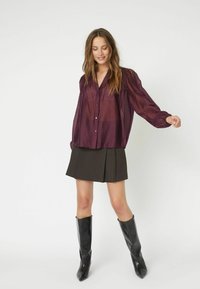 Camicia bordeaux con bottoni e maniche lunghe a sbuffo, abbinata a una gonna nera plissettata e stivali neri lucidi al ginocchio.