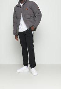 Grå pufferjakke med logo, hvid t-shirt, sorte sweatpants med sideløbende striber og hvide sneakers. Glat tekstur, afslappet pasform.