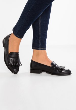 Mocassins en cuir noir avec un talon plat, ornés de franges et d'accents métalliques, présentés à côté de pantalons en denim foncé.