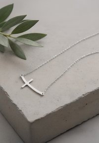 Srebrny naszyjnik krzyżykowy o eleganckim, minimalistycznym designie, z gładką powierzchnią i owalnym łańcuchem, wyeksponowany na teksturowanej szarej powierzchni.