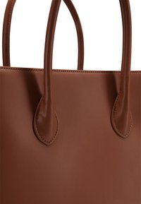 Brązowa skórzana torebka z dwiema okrągłymi rączkami i widocznym szwem w pobliżu mocowania rączek na gładkim tle.