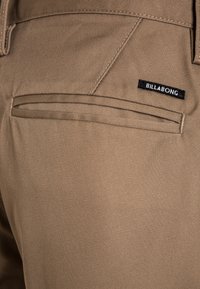 Kieszonka w khaki spodniach z poziomym rozcięciem i małą czarną metką z napisem "BILLABONG" wszytą nad kieszonką.