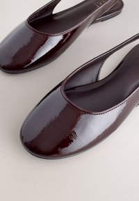Paire de ballerines à bride arrière en cuir verni brun foncé brillant avec bouts arrondis sur une surface claire.