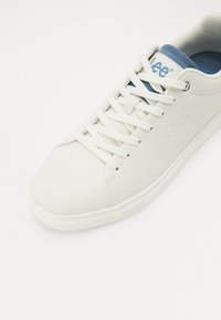 Baskets blanches avec lacets blancs, doublure intérieure bleue, et logo de la marque Lee embouti sur le côté et imprimé sur la languette.